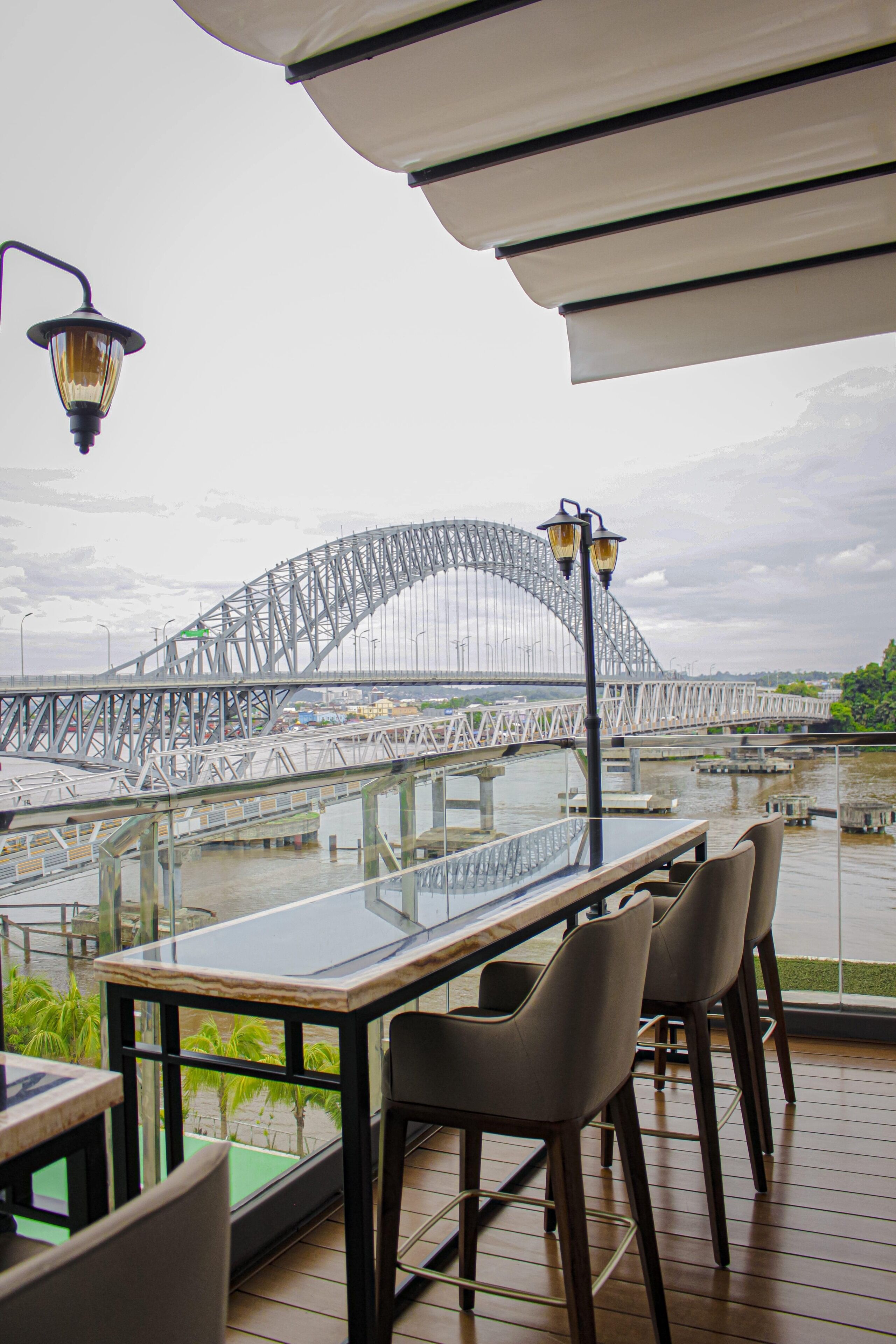 A restaurant with a view of a large bridge, outdoor seating, and a glass table.