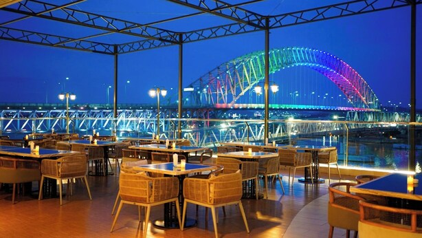 Un área de comedor al aire libre con mesas y sillas, vista a un puente iluminado y una barandilla con vista a un cuerpo de agua.