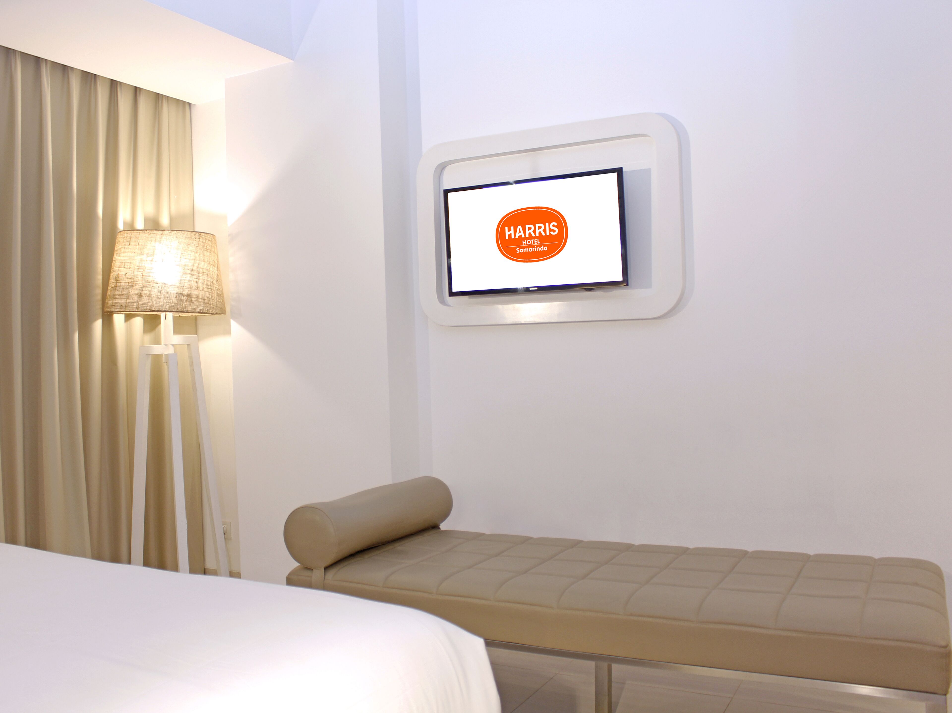 A hotel room with a bed, a bedside table, a lamp, and a digital display showing "HARRIS HOTEL Samarinda".