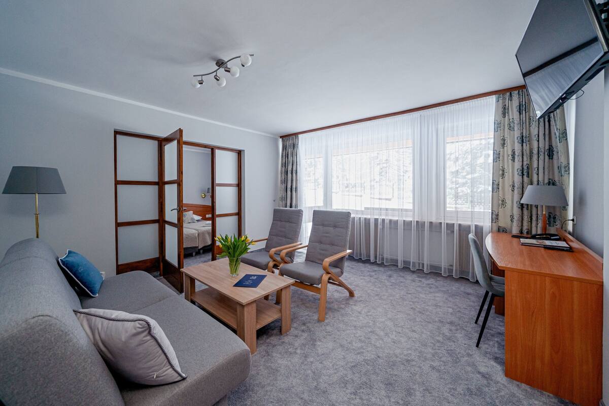 Een moderne hotelkamer met een grijze bank, een houten bureau, twee fauteuils, een klein tafeltje met een plant en een groot raam met dunne gordijnen.