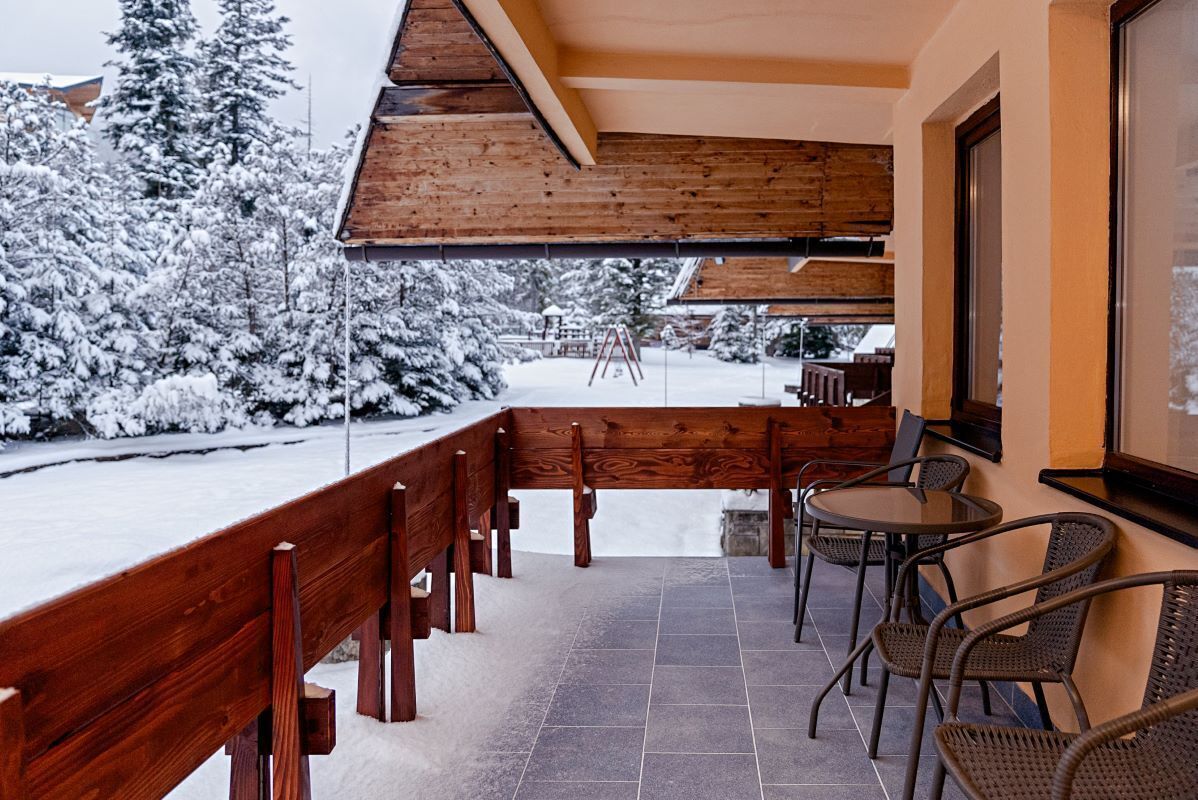 A covered outdoor seating area with a wooden bench and chairs, a tiled floor, and snow-covered trees in the background.