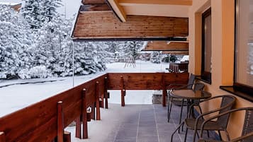 A covered outdoor seating area with a wooden bench and chairs, a tiled floor, and snow-covered trees in the background.