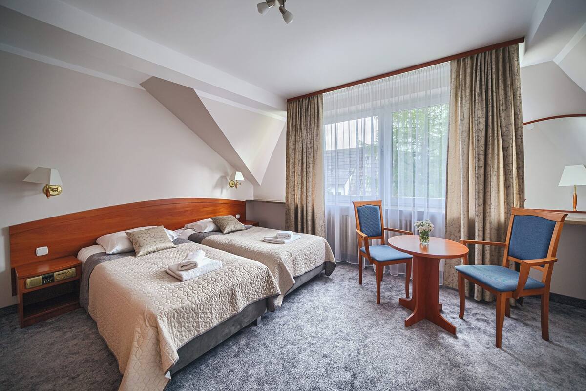 Een hotelkamer met twee bedden, een klein tafeltje met stoelen, en een groot raam met gordijnen.