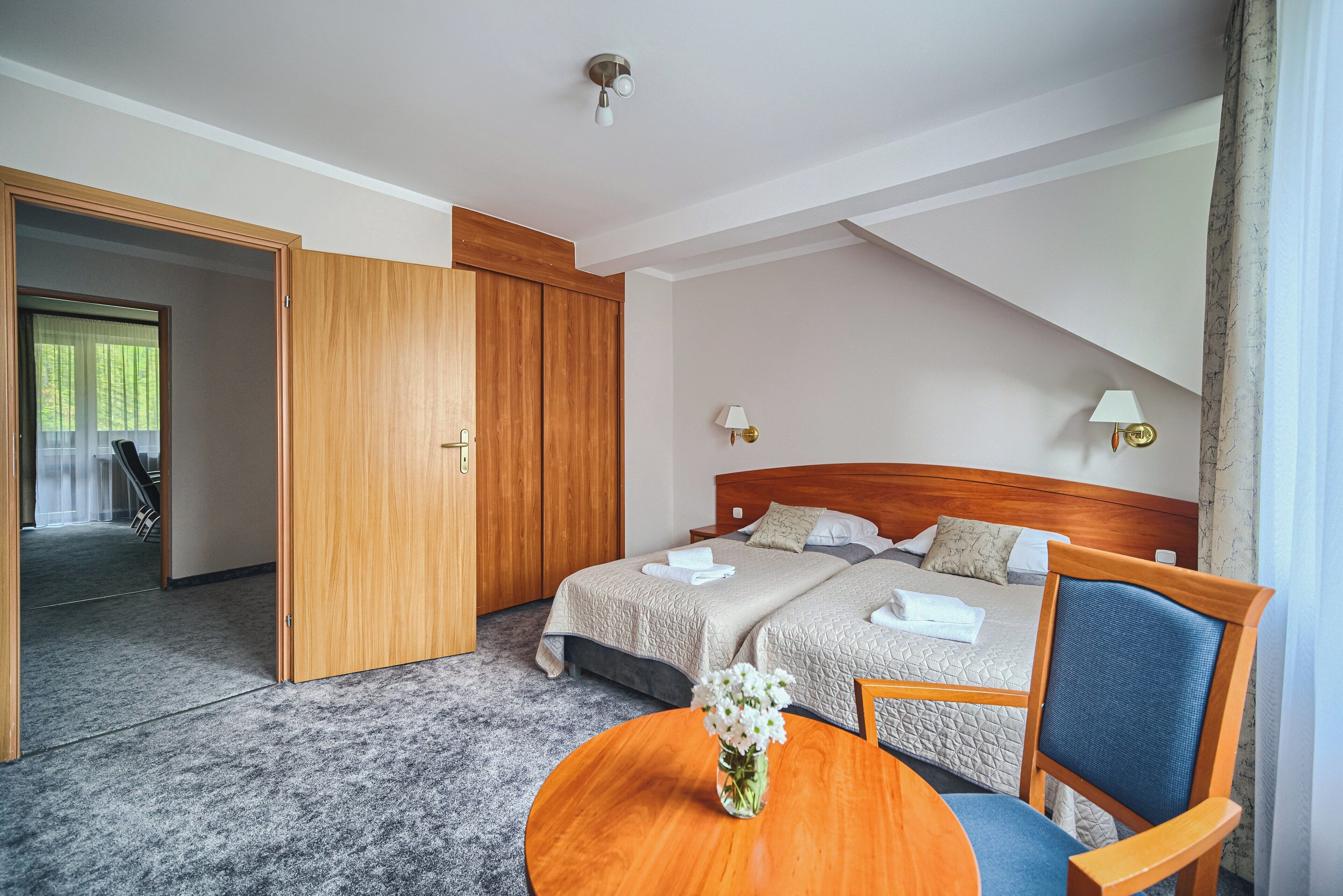 A hotel room with two beds, a wooden wardrobe, a round table with a vase, and a chair.