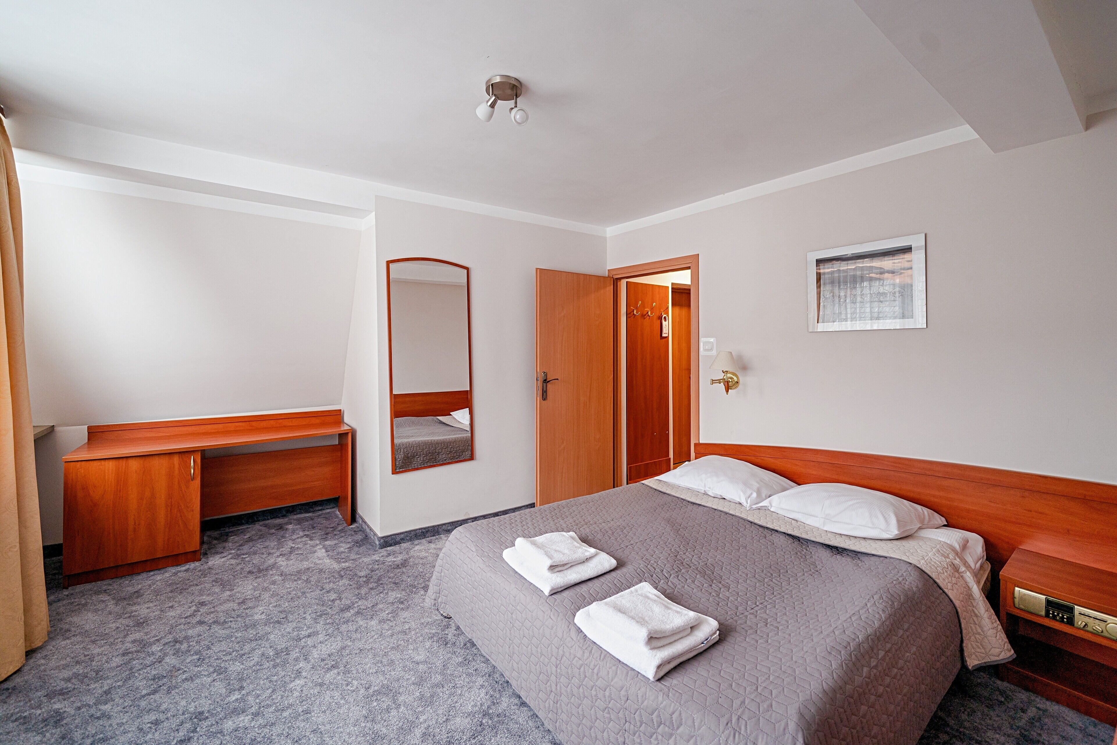 A hotel room with a bed, a desk, a mirror, a door, and a nightstand.