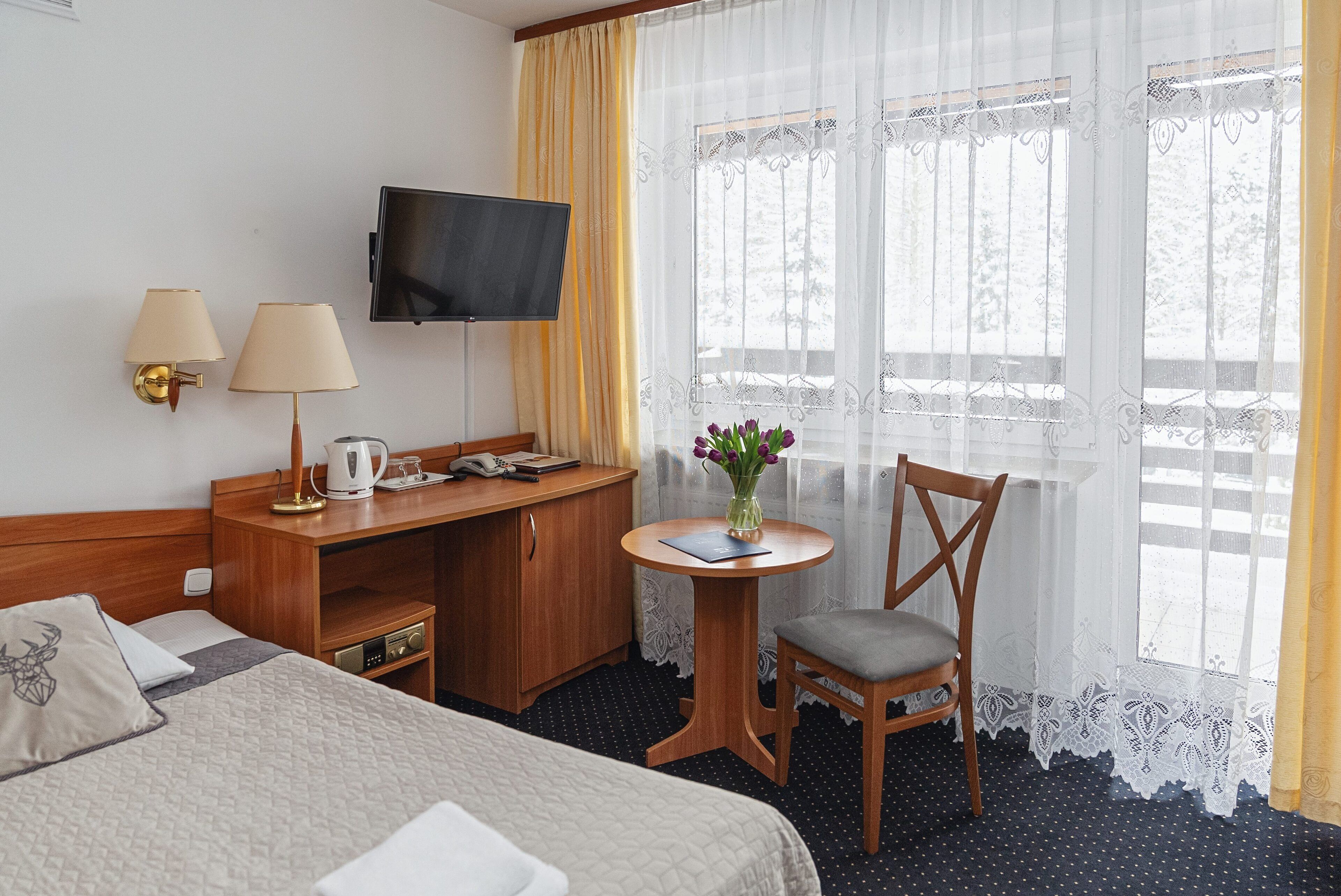 Habitación de hotel con cama, escritorio y silla, televisor, una mesita con un jarrón de flores y ventana con cortinas.