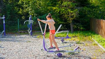 A child using an outdoor exercise machine in a playground with other fitness equipment and a fence in the background.