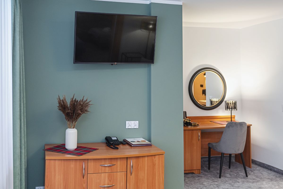 A hotel room with a flat-screen TV mounted on the wall, a wooden desk with a chair, a round mirror, and a vase with dried plants on the dresser.
