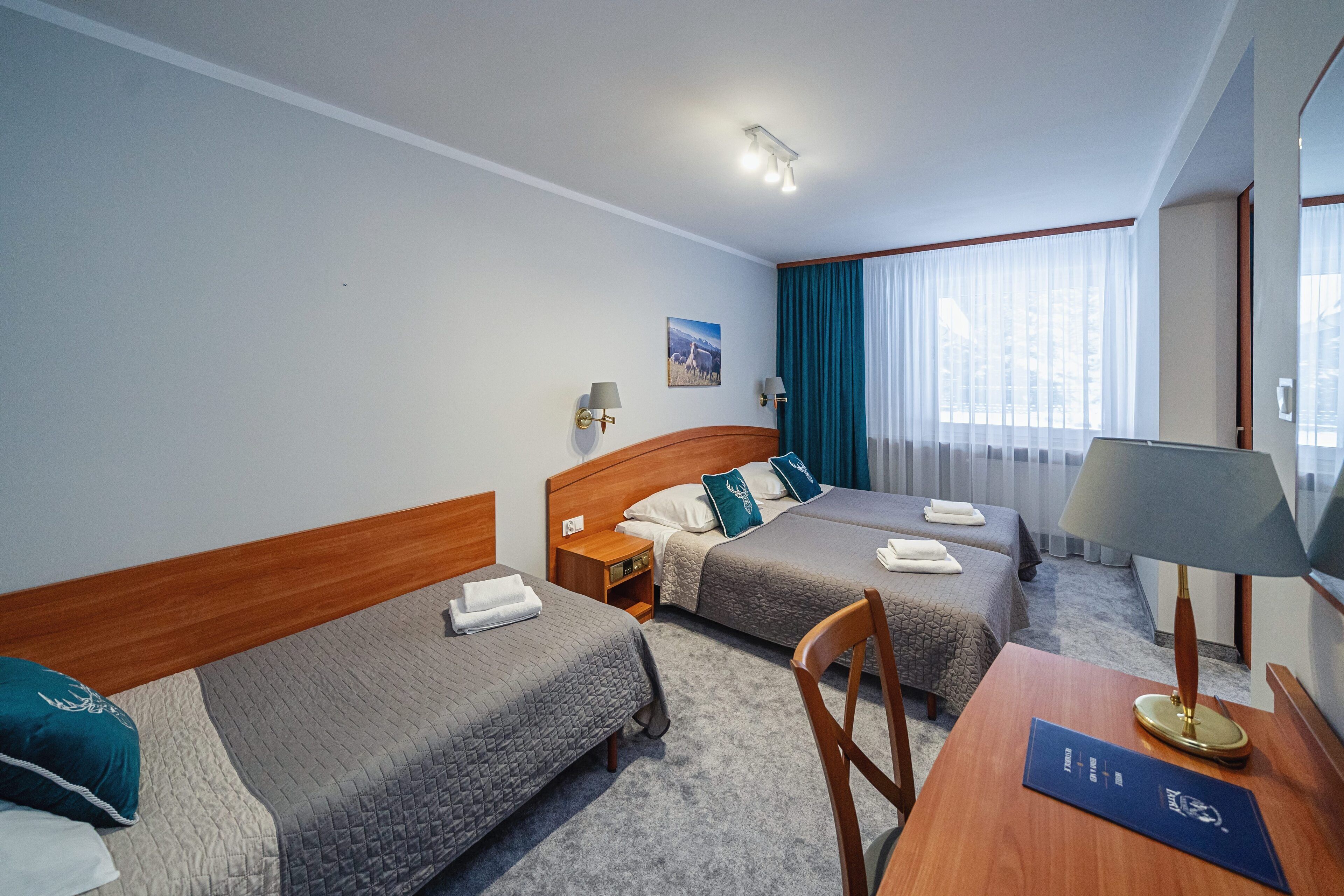 Habitación de hotel con dos camas individuales, un escritorio de madera, una silla, una lámpara, una mesita de noche y una ventana con cortinas.