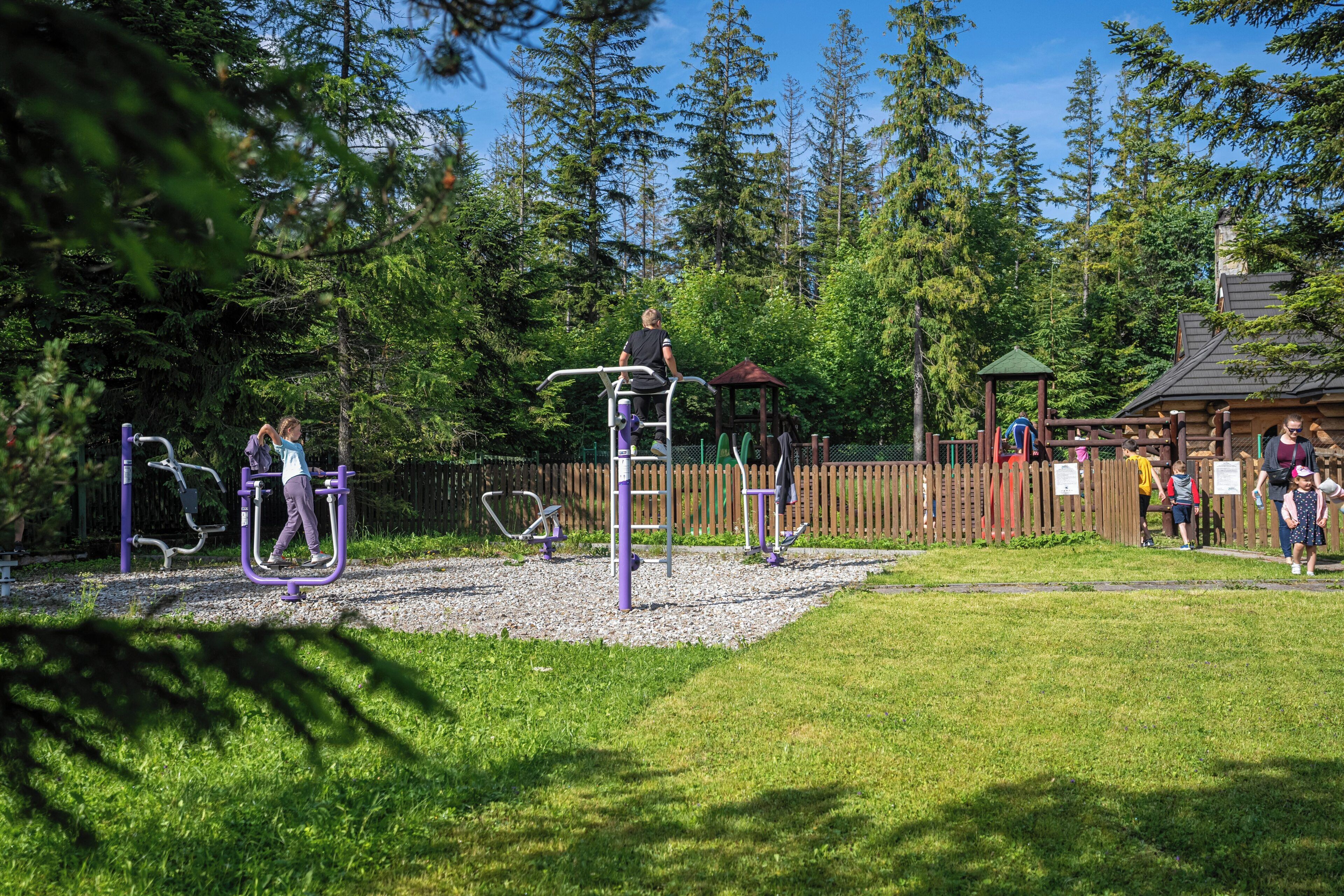 An outdoor playground with exercise equipment, surrounded by trees and a wooden fence.