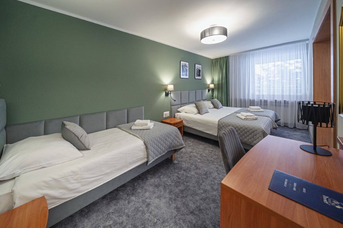Een hotelkamer met twee eenpersoonsbedden, een bureau en een stoel. Er zijn twee nachtkastjes met lampen, een groot raam met gordijnen en een plafondlamp.