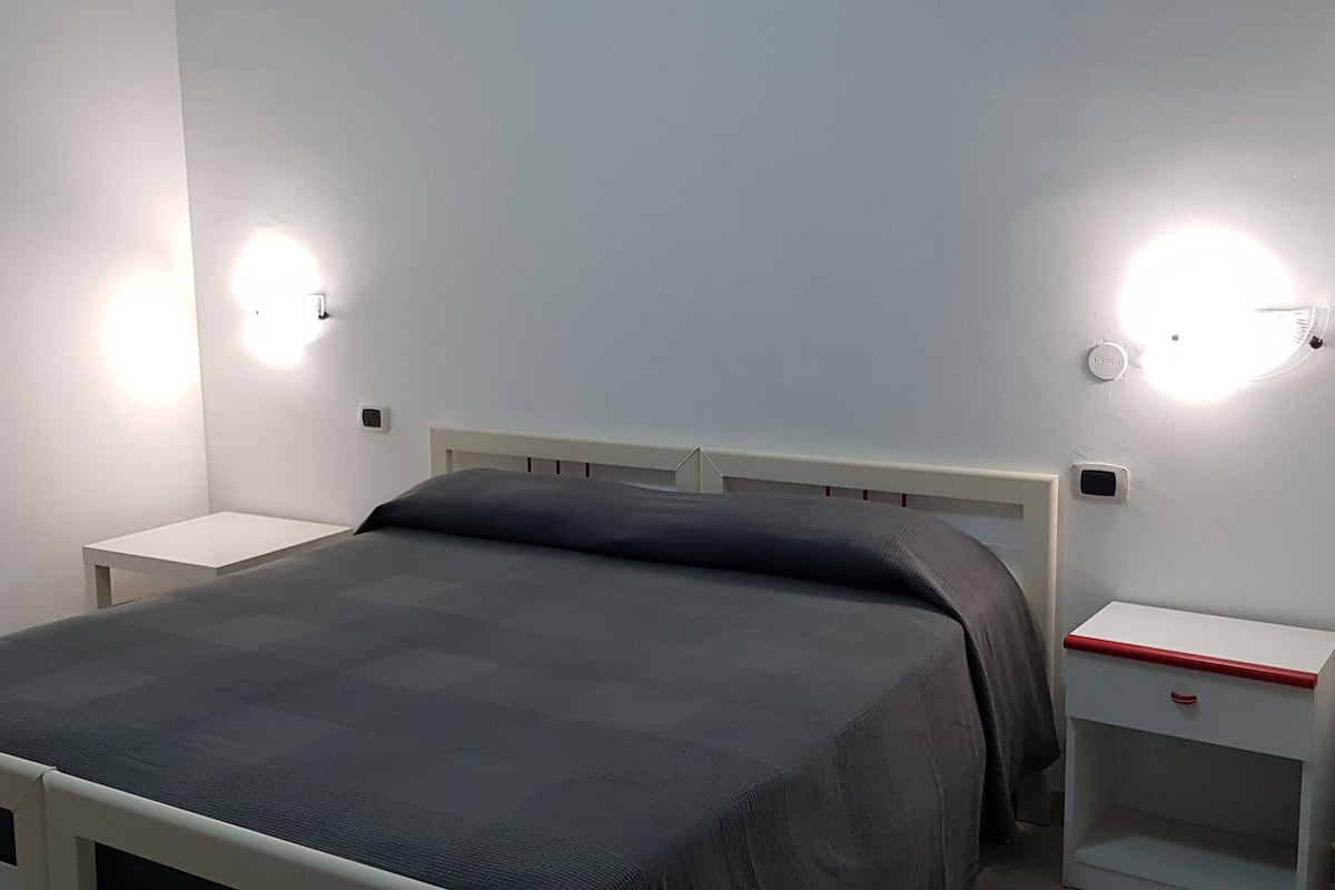 A single bed with a grey mattress and white headboard in a room with white walls and two bedside tables with red handles.