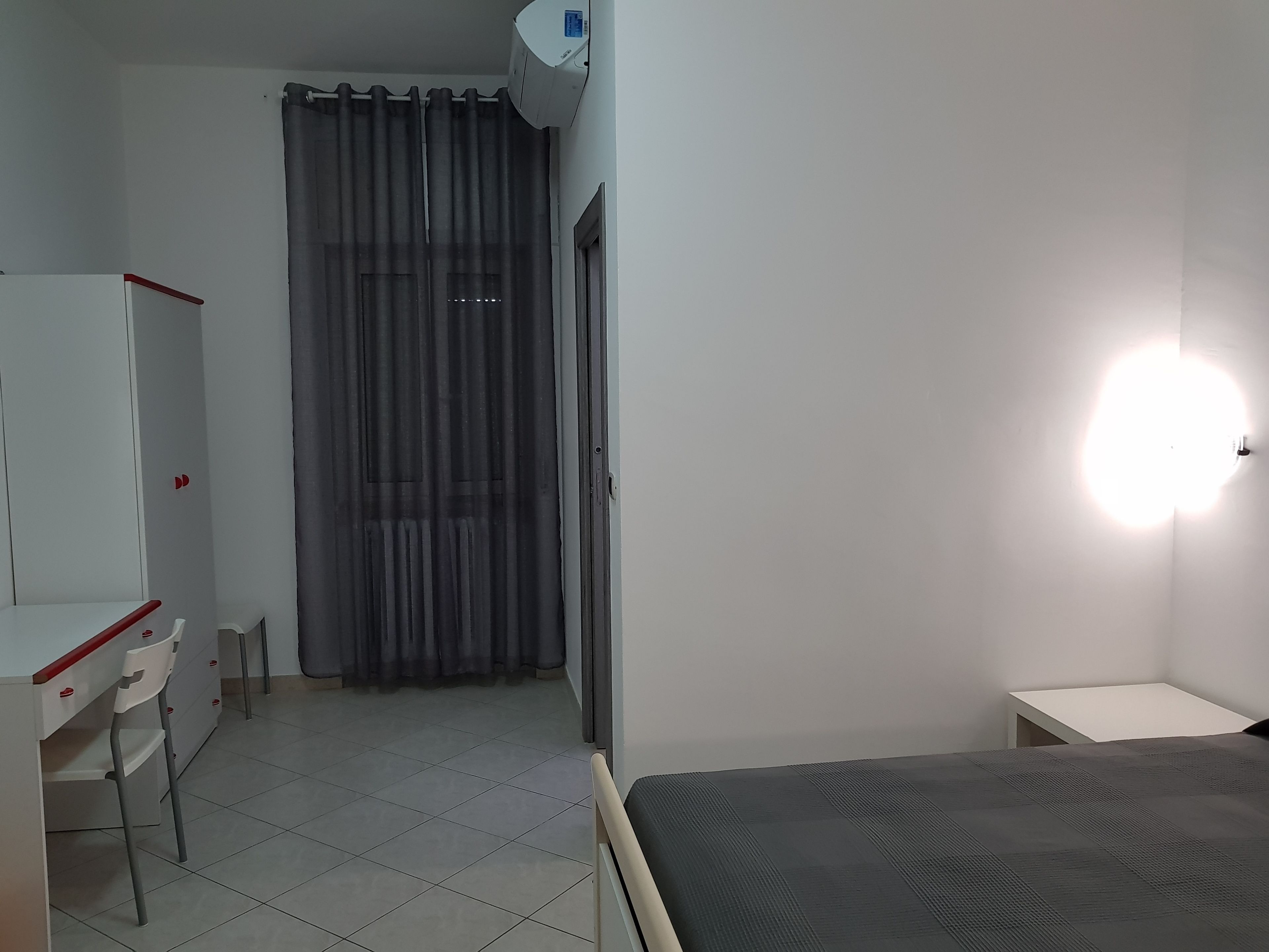 A room with a bed, a desk, and a chair. There is a curtain, a wall-mounted air conditioner, and a light fixture.