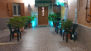Un pasillo con piso estampado, paredes de madera y una puerta con la etiqueta "Holiday B&B". Se observan plantas en macetas y sillas a lo largo de los lados.