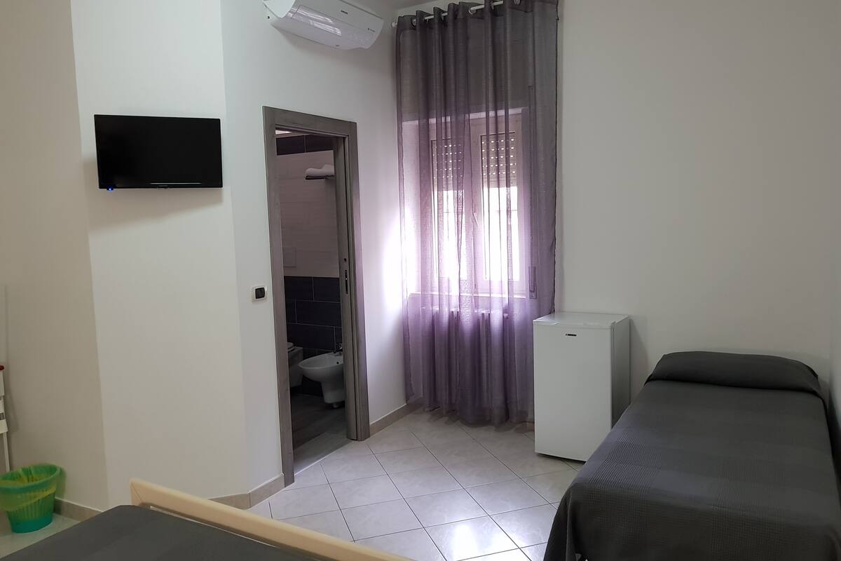 A small, clean room with a bed, a TV mounted on the wall, a refrigerator, and a bathroom visible through a doorway.