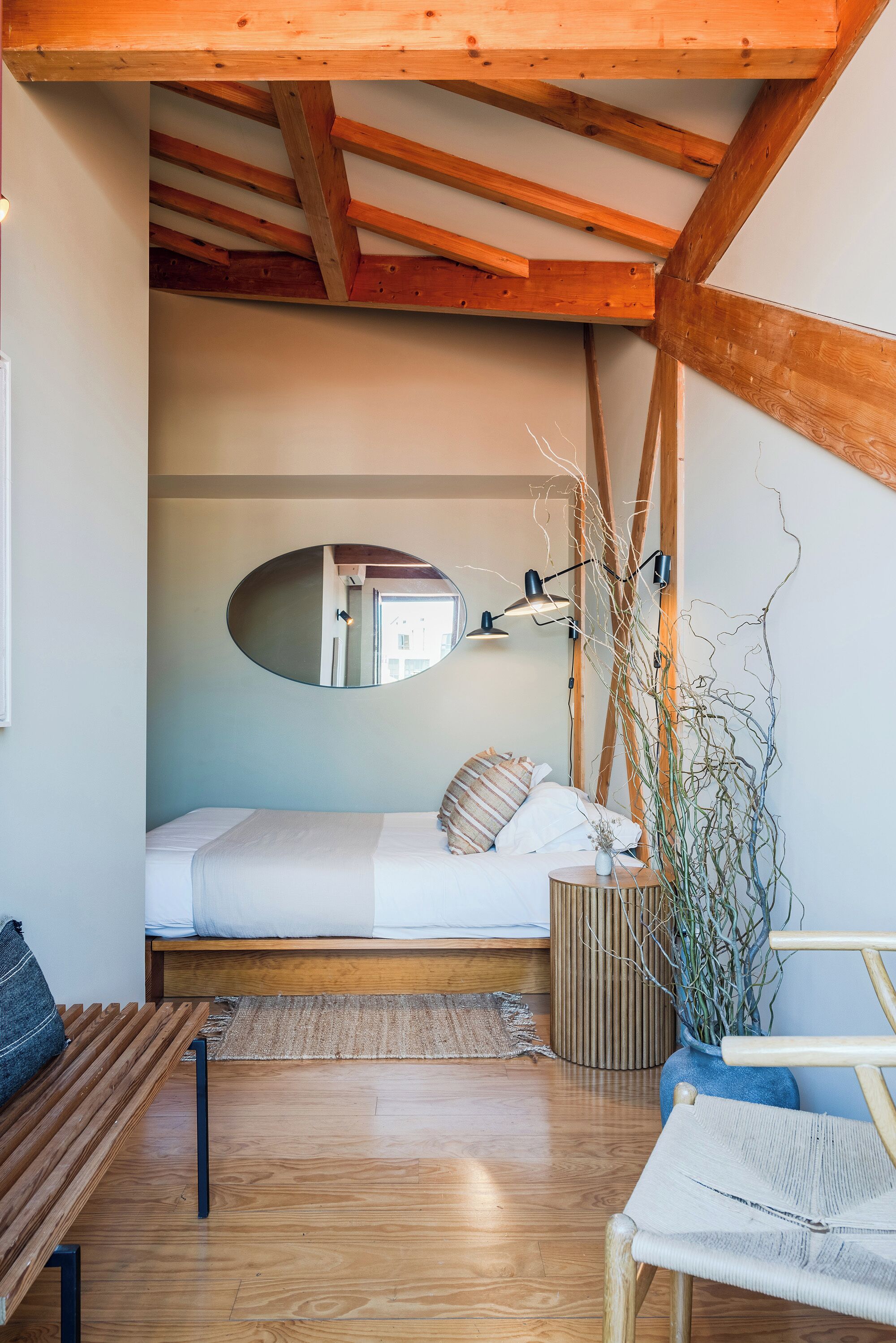 A compact bedroom with a wooden ceiling, a single bed, a bedside table with a lamp, a mirror, and a potted plant.