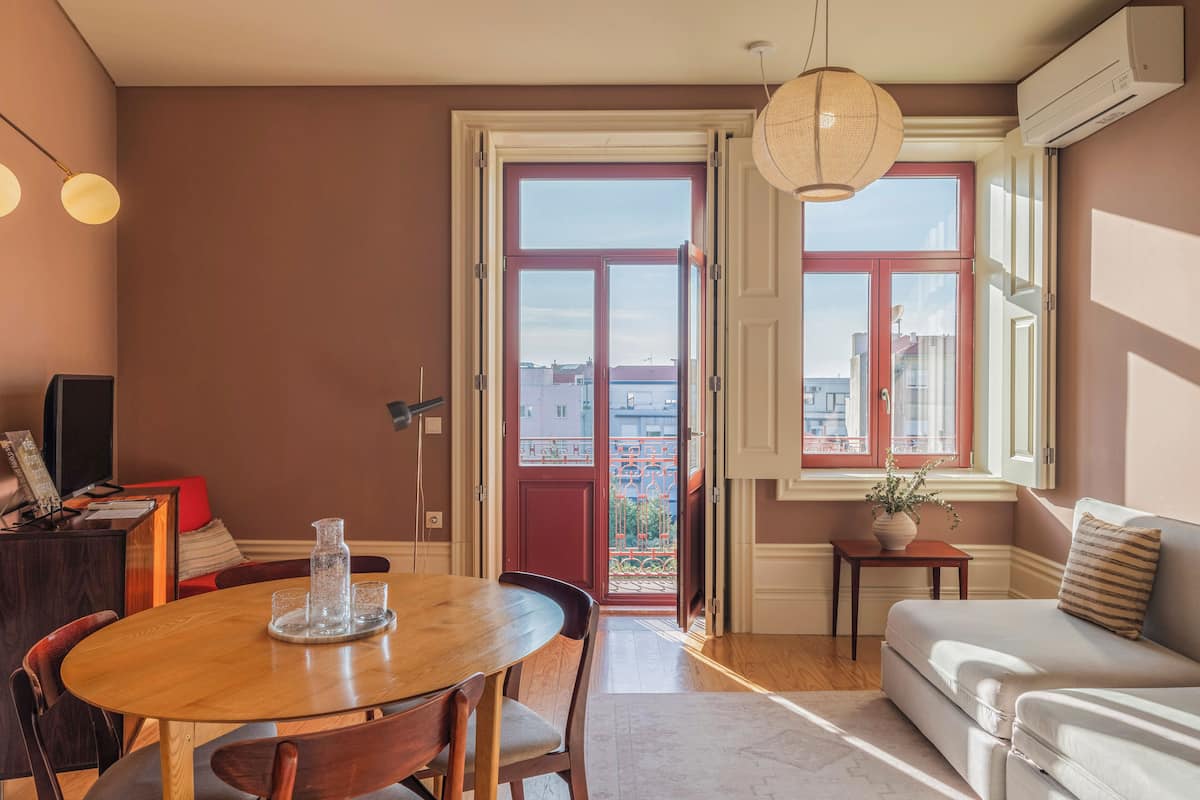 A cozy living room with a wooden dining table, a sofa, and a view through a red-framed door to the outside.