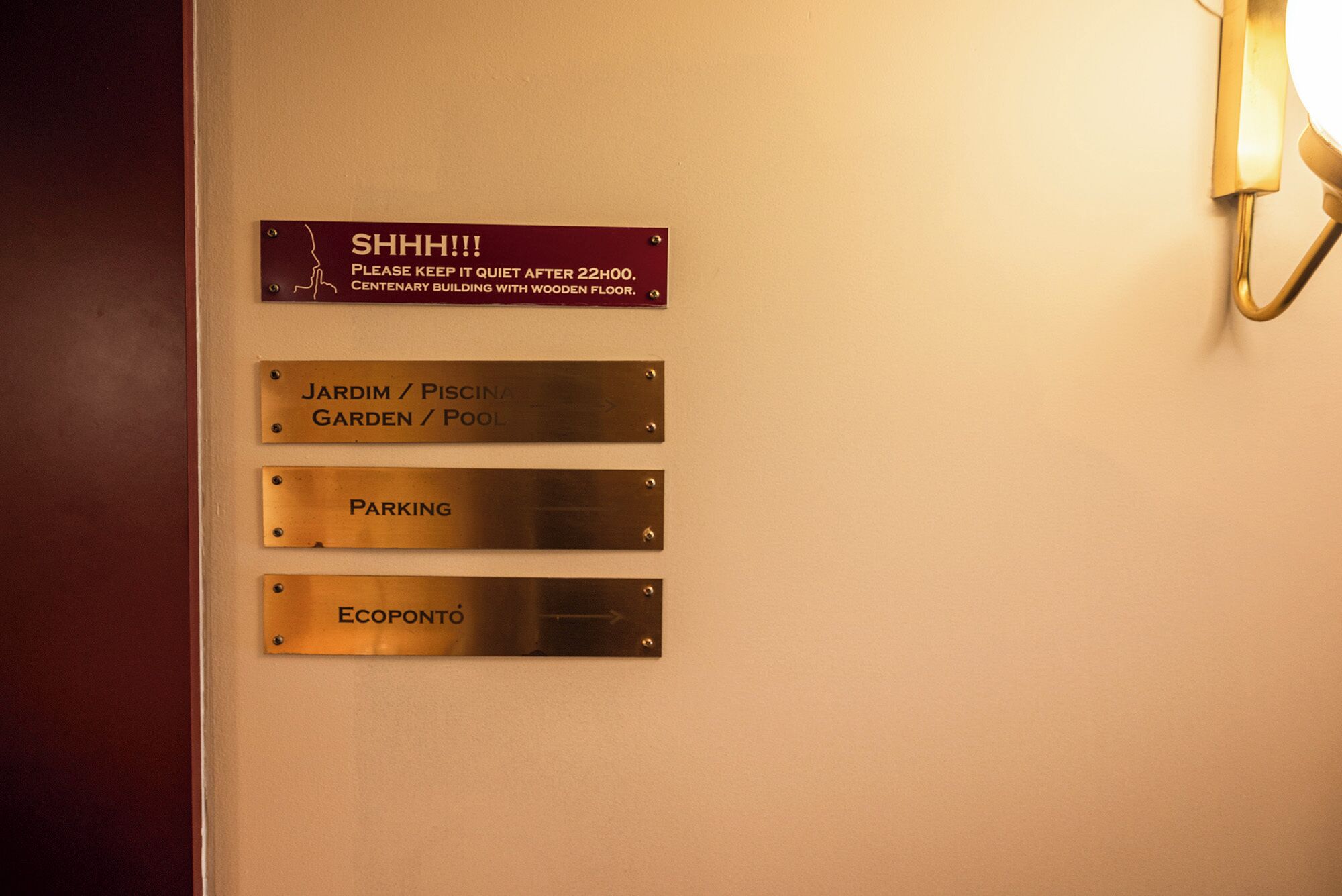 A wall with directional signs for 'SHHH!!! PLEASE KEEP IT QUIET AFTER 22:00. CENTENARY BUILDING WITH WOODEN FLOOR.', 'JARDIM / PISCINA GARDEN / POOL', 'PARKING', and 'ECOPONTO'.