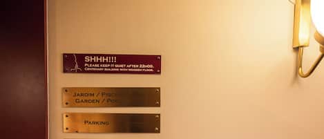 A wall with directional signs for 'SHHH!!! PLEASE KEEP IT QUIET AFTER 22:00. CENTENARY BUILDING WITH WOODEN FLOOR.', 'JARDIM / PISCINA GARDEN / POOL', 'PARKING', and 'ECOPONTO'.