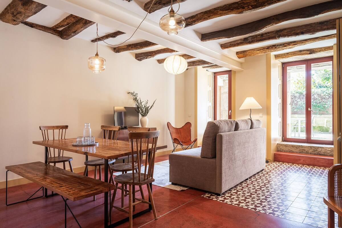A dining area with a wooden table, chairs, and a sofa. There is a TV, a vase with plants, and a lamp. The room has exposed wooden beams on the ceiling and a window with a view of greenery.