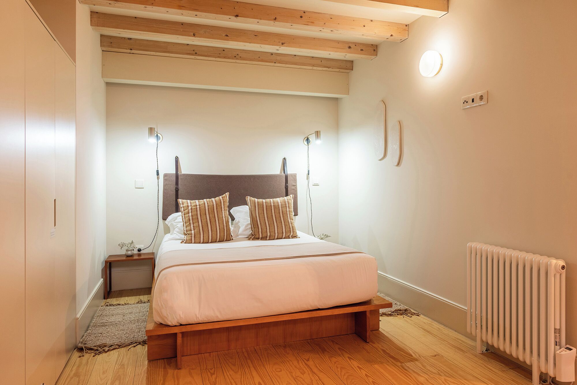 Un dormitorio con estructura de cama de madera, una mesita de noche, una lámpara de noche y un aplique de pared.