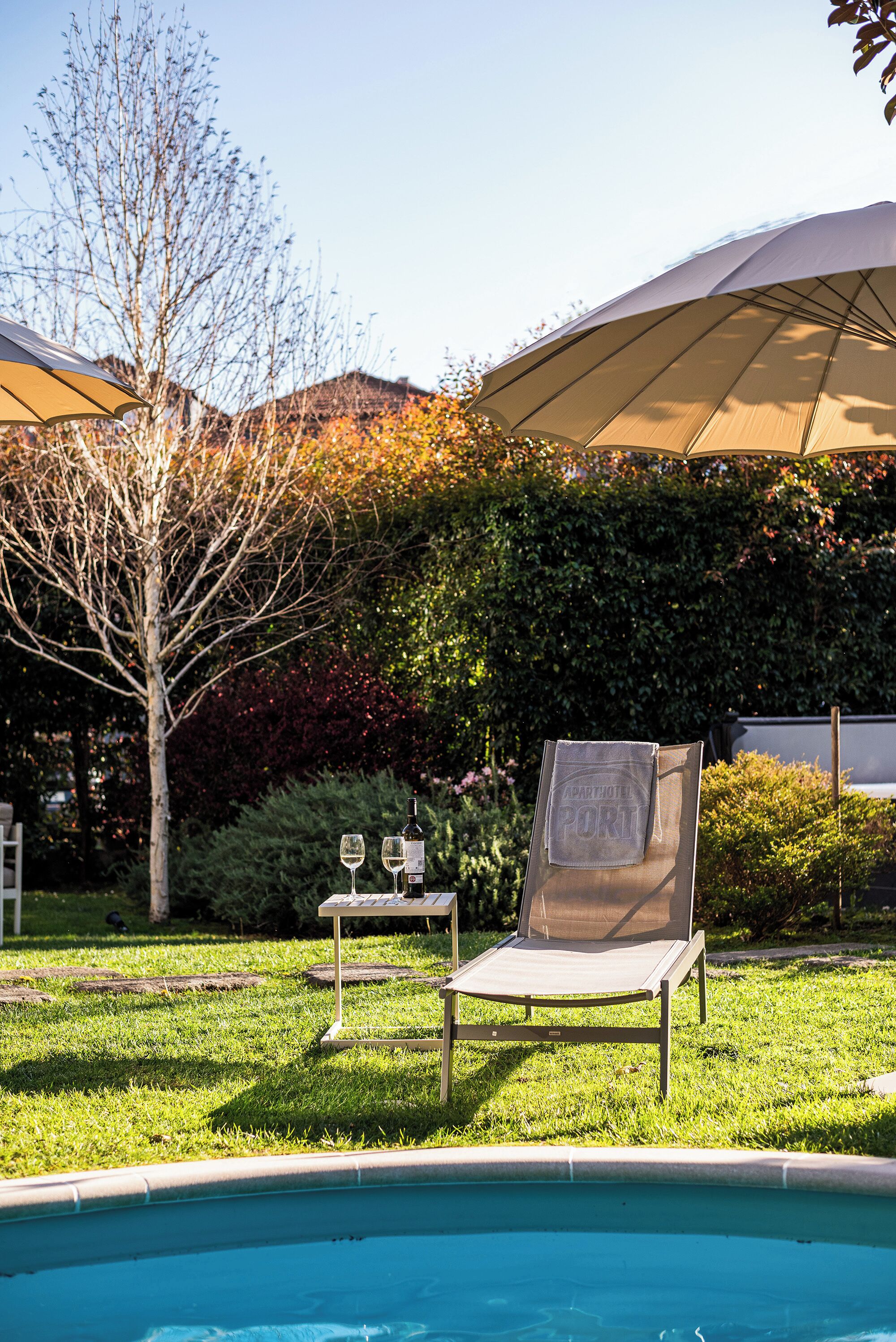 A poolside area with a lounge chair, a table with a bottle and glasses, and a large umbrella.