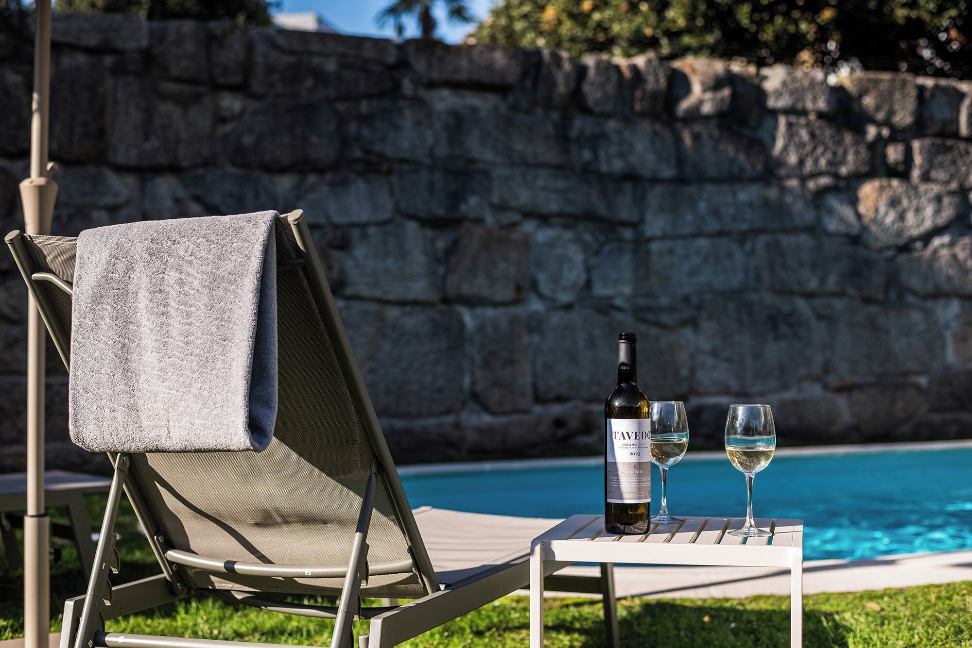 Una silla de descanso junto a la piscina con una toalla, una botella de vino y dos copas sobre una mesita.