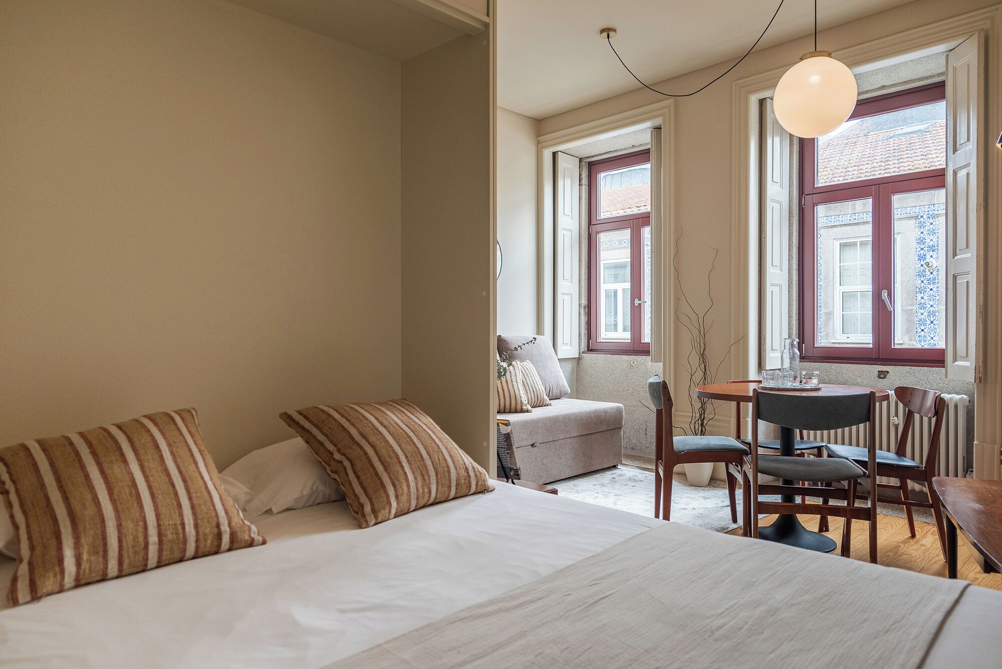 Quarto com cama, uma mesinha e duas cadeiras. Janelas com molduras vermelhas e vista para o exterior.