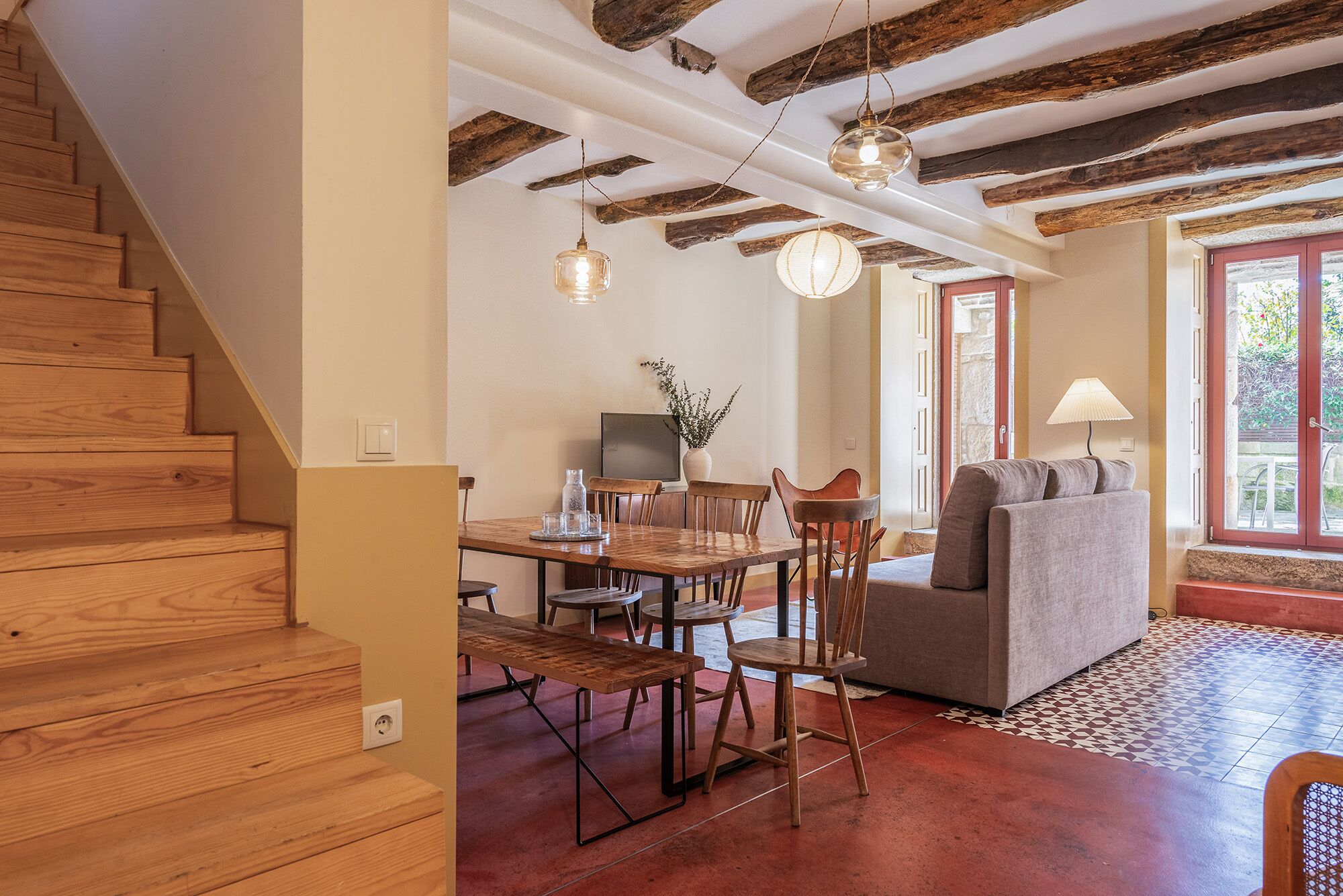 A spacious living area with a wooden staircase, a dining table with chairs, a gray sofa, and a TV.