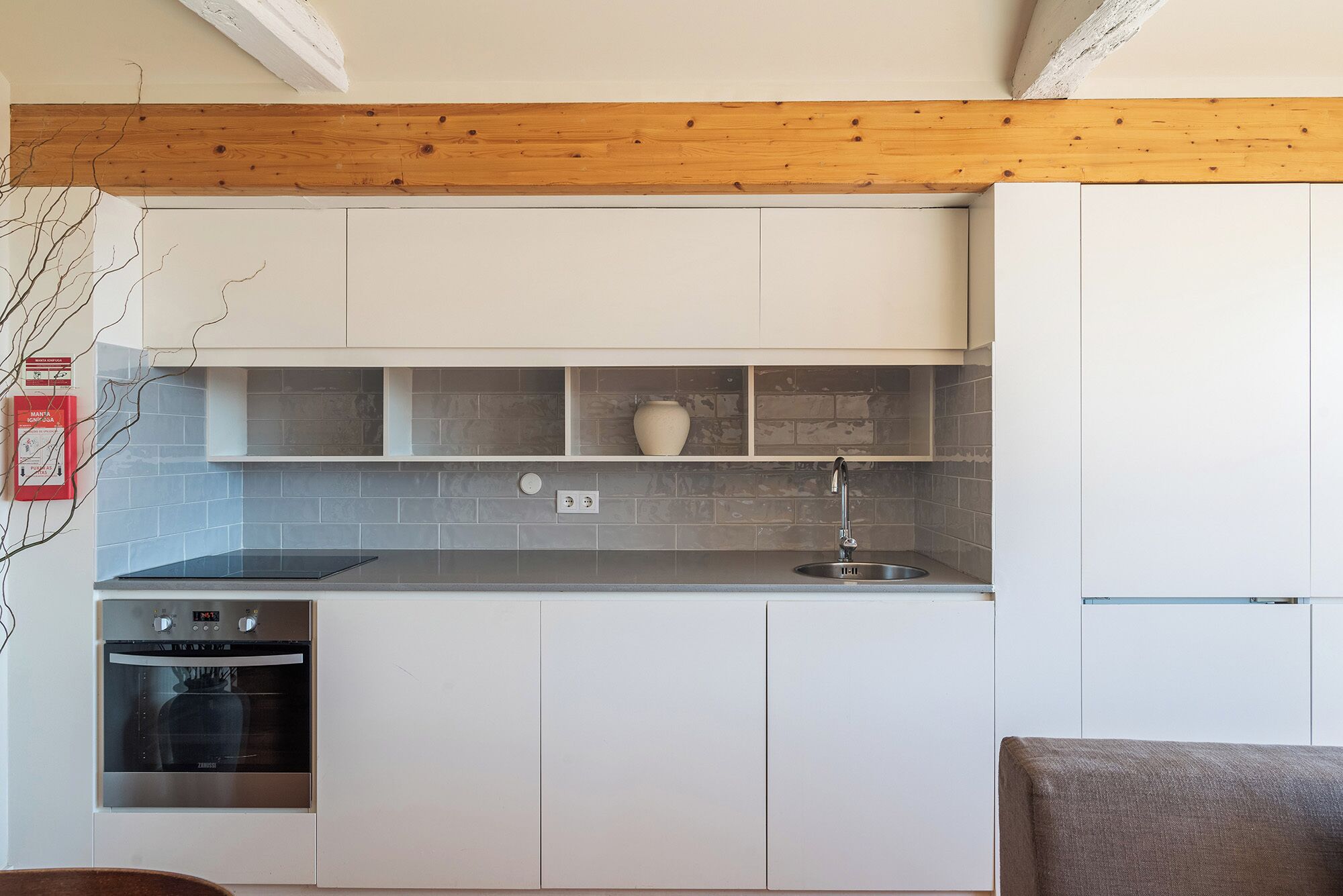 A modern kitchen with white cabinets, a grey tiled backsplash, a stainless steel oven, and a wooden ceiling beam.