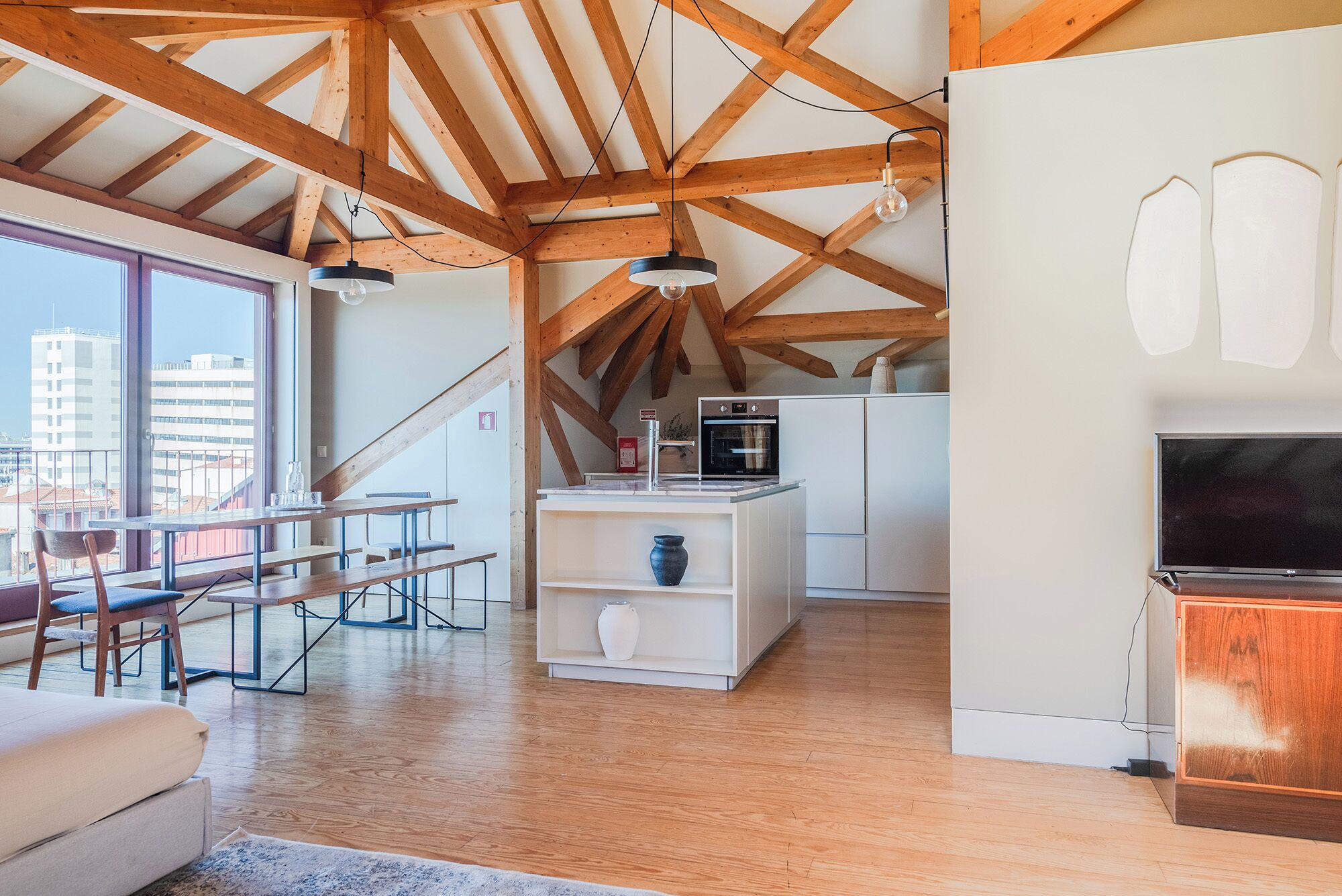 A spacious loft with high ceilings, exposed wooden beams, a dining area with a table and chairs, a kitchen with modern appliances, and a large window offering a city view.