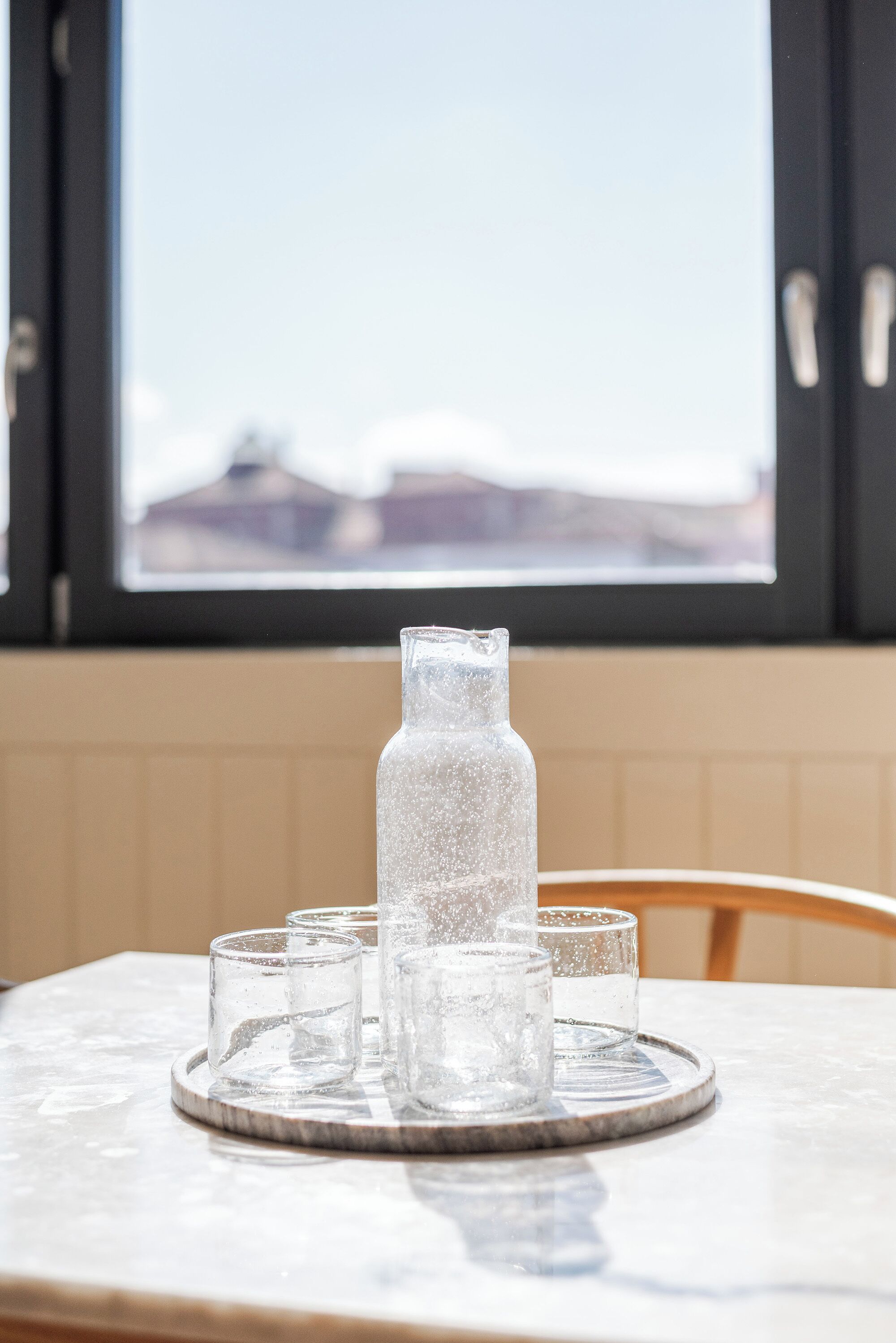Et bord med en klar glasflaske og fire glas på et fad, placeret foran et vindue med udsigt til bygninger.