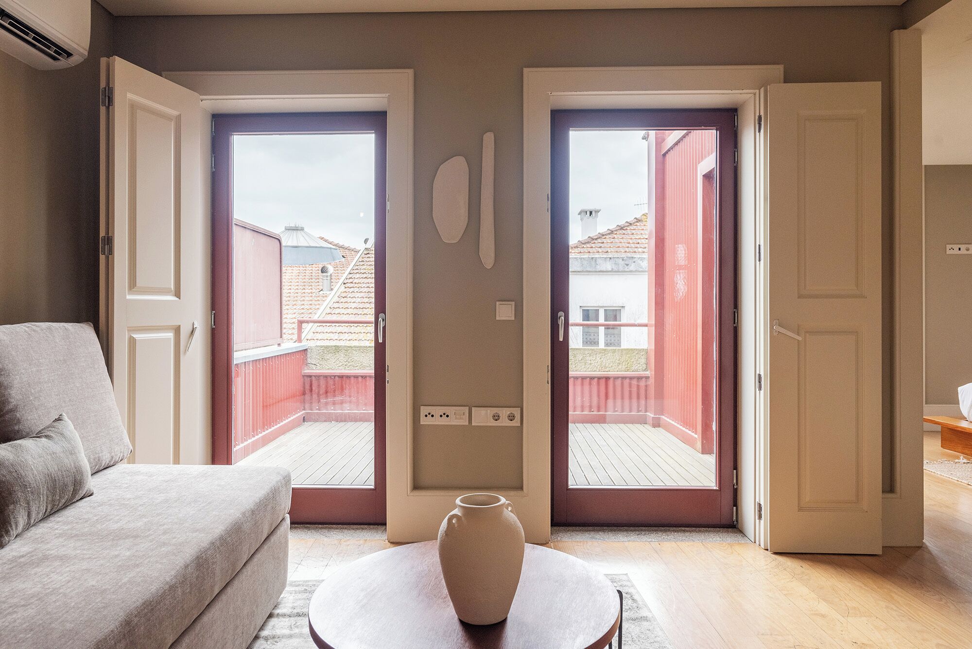 A room with a grey sofa, a round table with a vase, and two open sliding doors leading to a balcony with red railings.