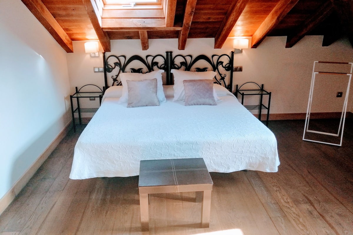 A bedroom with a wooden ceiling, a bed with white bedding, two bedside tables, and a bench.