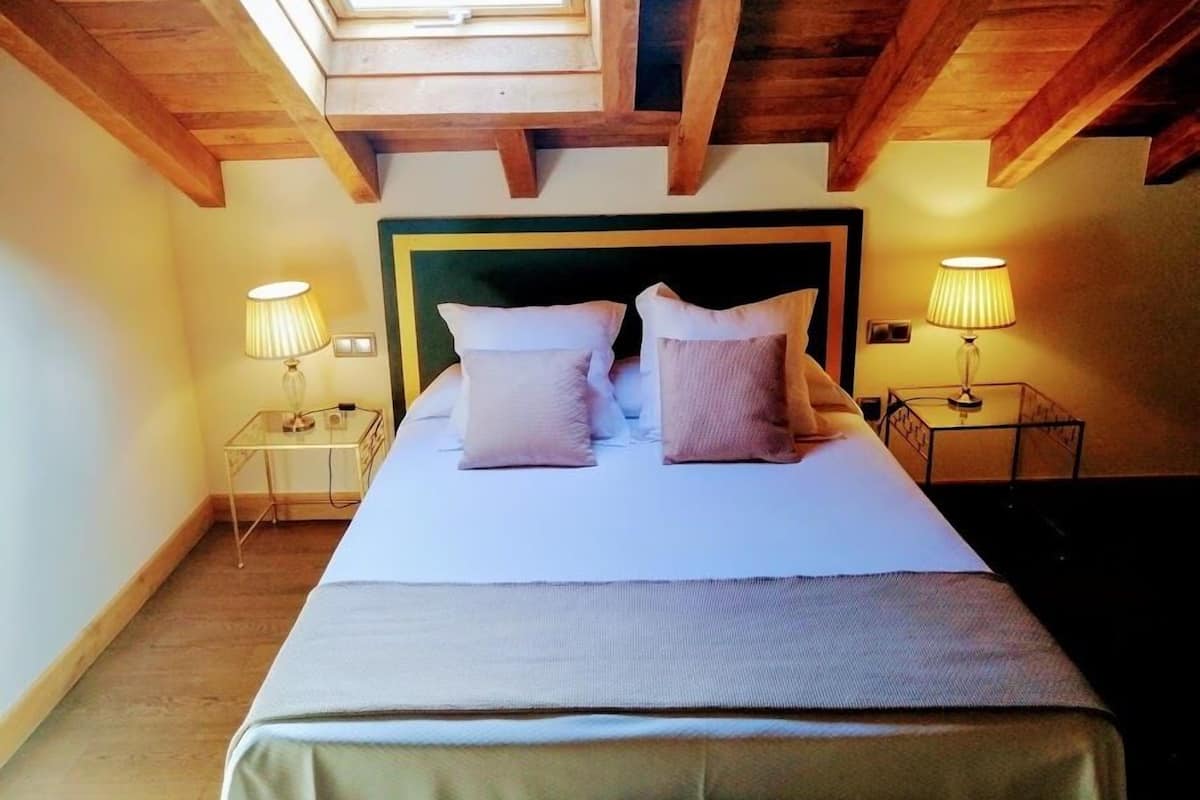 A bedroom with a skylight, wooden beams, a bed with pillows, and bedside lamps.
