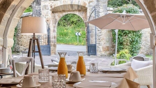 An outdoor dining area with stone arches, a table set for two, a lamp, and a view of a stone bridge and greenery. - Hotel Palacio de Arce (Pielagos)