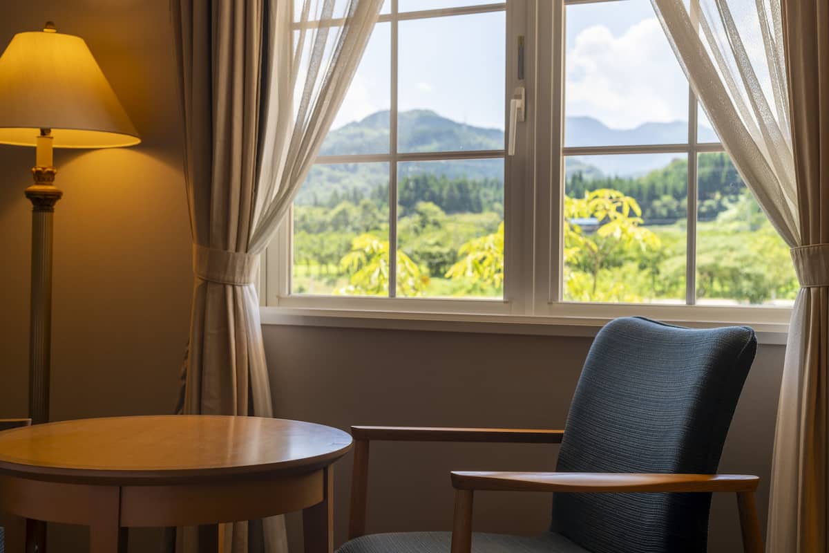A room with a view of a mountainous landscape through a window, a chair, a table, and a lamp.