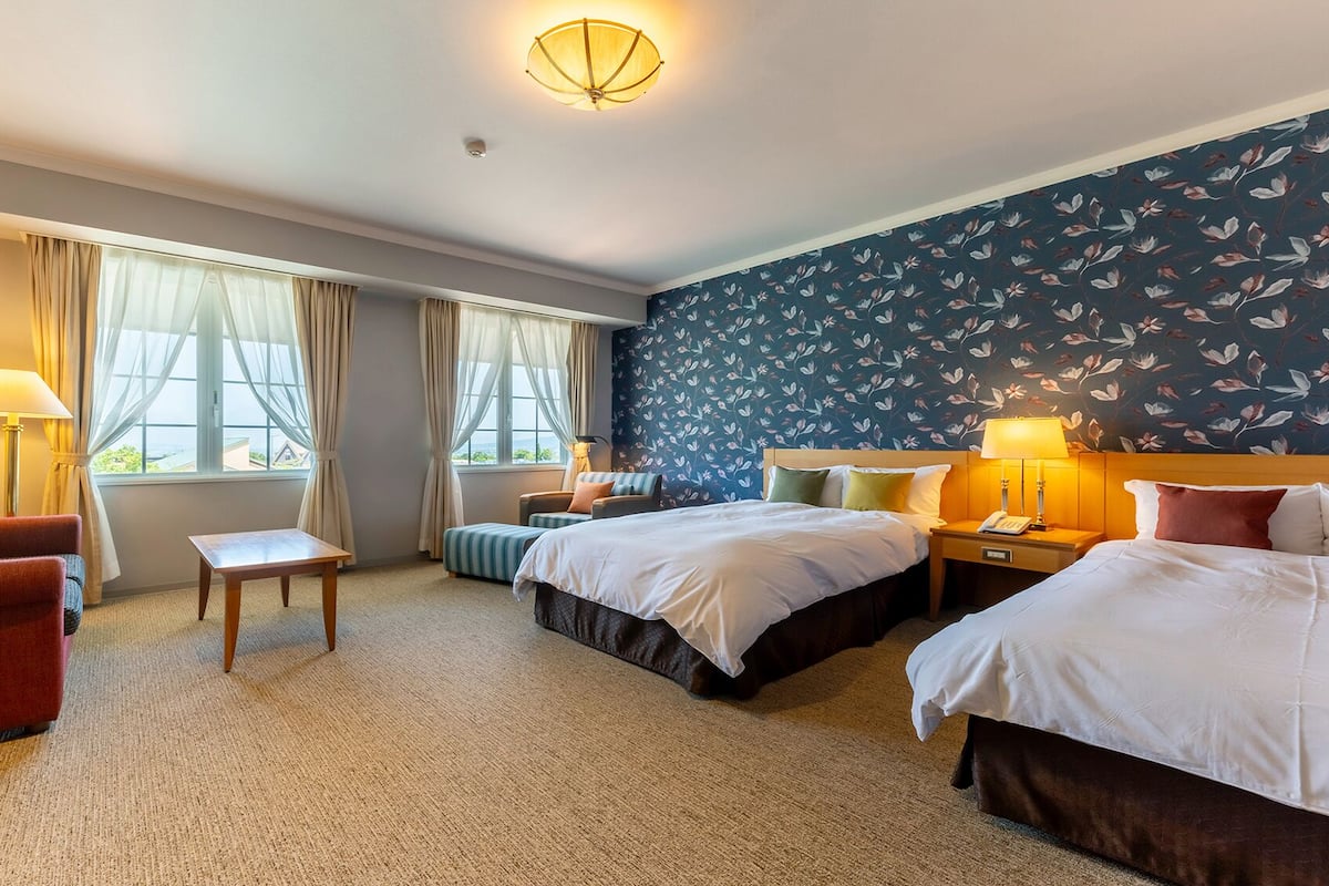 A hotel room with two beds, a small table, a chair, and a sofa. There are curtains on the windows, and the room has a carpeted floor.
