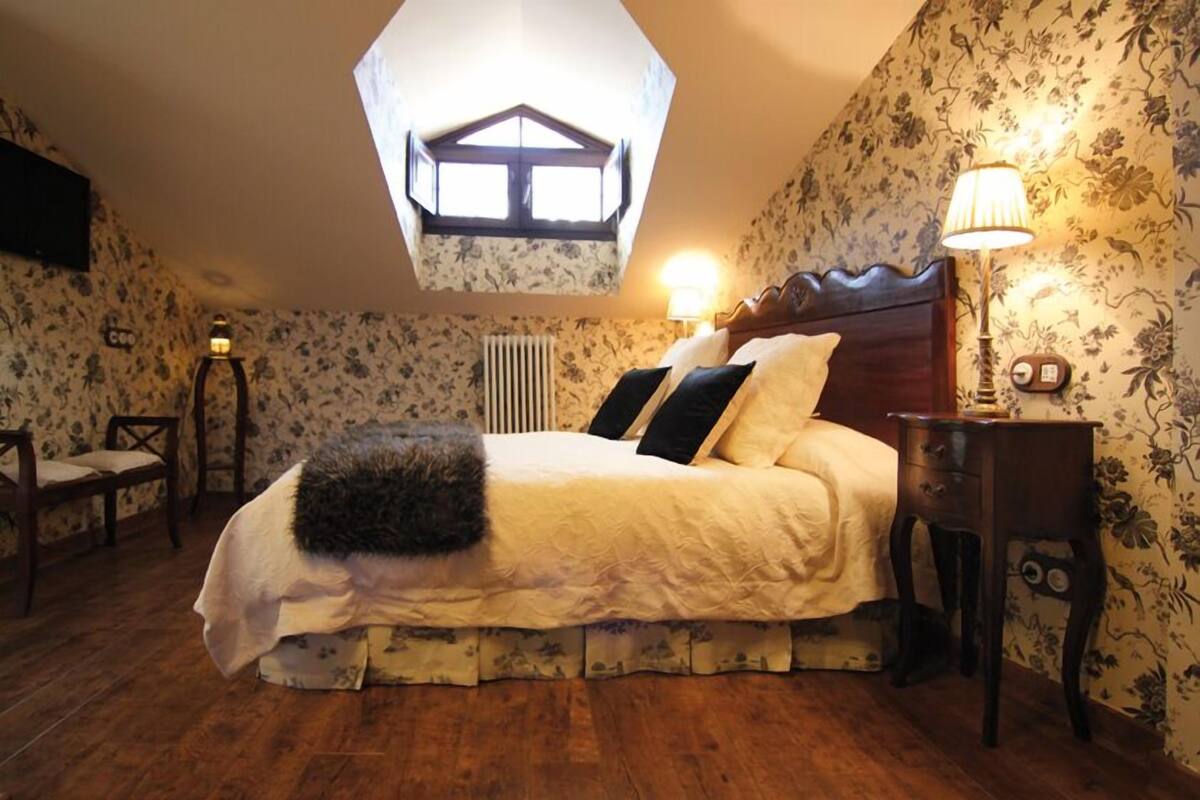 A bedroom with a large bed, a wooden headboard, a skylight, a nightstand, and floral wallpaper.