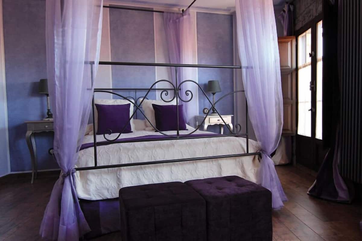 A bedroom with a four-poster bed, a nightstand, a lamp, and a window with curtains.