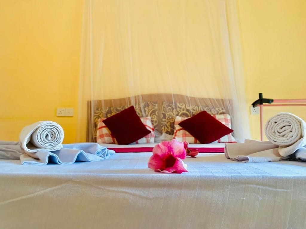 Un letto rifatto con una coperta bianca, due asciugamani arrotolati, un fiore rosa e una tenda a baldacchino.