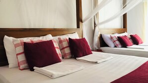 Una cama bien hecha con sábanas blancas y almohadas rojas y blancas, un cabecero de madera y una ventana con cortinas de gasa.
