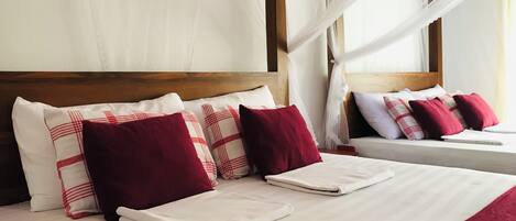 A neatly made bed with white linens and red and white pillows, a wooden headboard, and a window with sheer curtains.