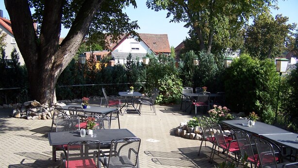 Een terras buiten met tafels en stoelen, omgeven door bomen en een bakstenen muur.