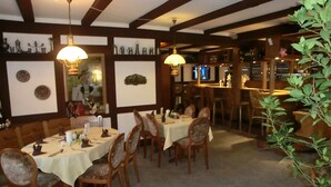 Een restaurant met houten balken, ronde tafels met witte tafellinnen en stoelen met beklede zitplaatsen.