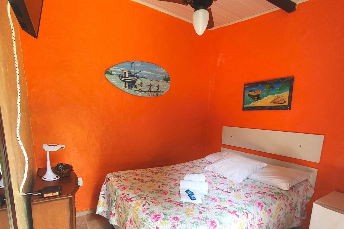 A bedroom with an orange wall, a bed with floral bedding, a ceiling fan, and a small table with a lamp.
