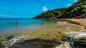 A beach with clear water, a person in the water, and a rocky shoreline. - Suites Casa do Barão (Búzios)