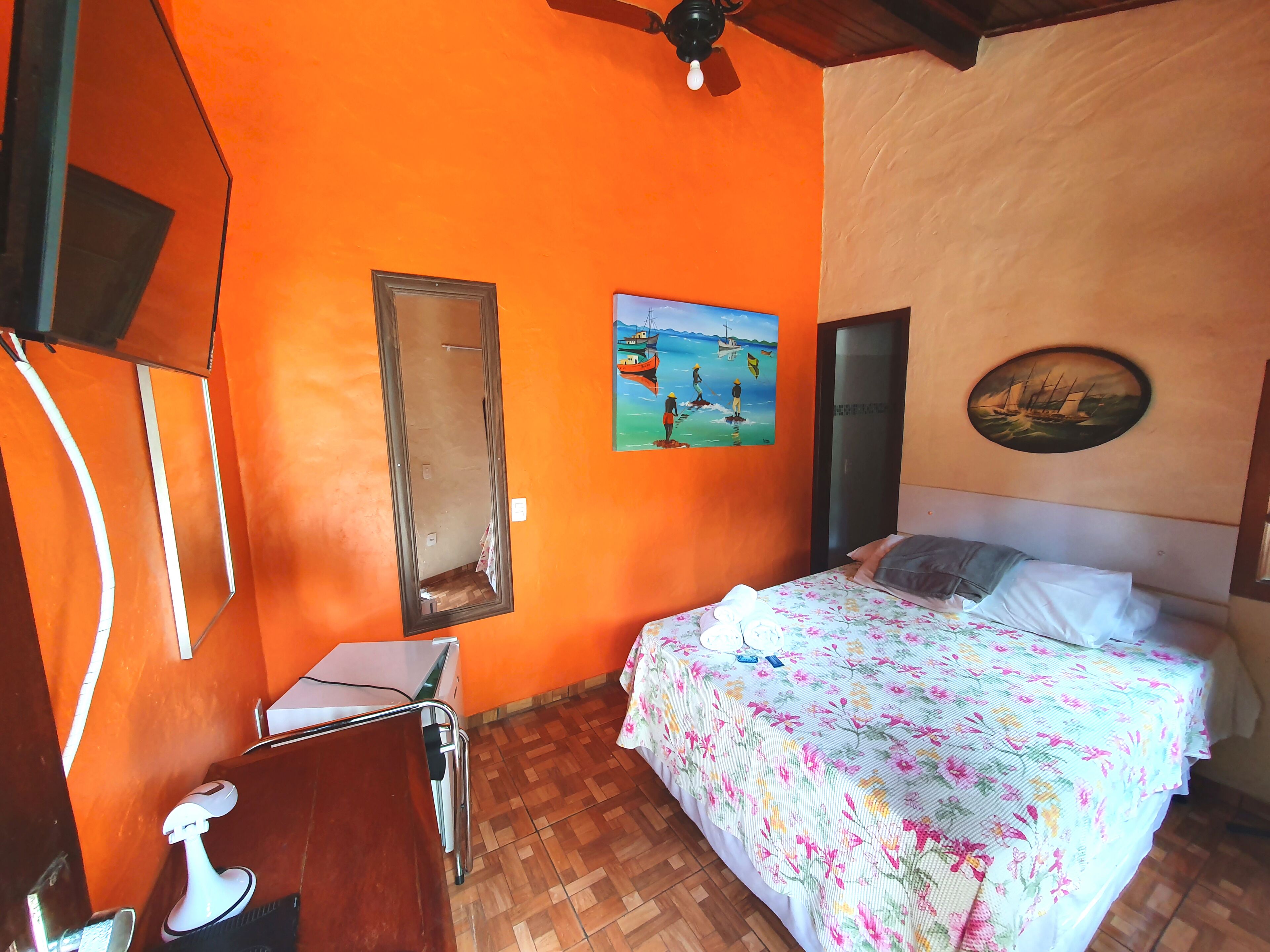 Una habitación con pared naranja, una cama con ropa de cama floral, un ventilador de techo y un cuadro de un paisaje marino.