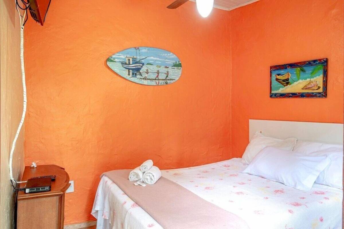 A bedroom with an orange wall, a bed with white linens, a painting of a boat, and a nightstand with a lamp.