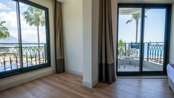 Ein Zimmer mit großen Fenstern, einem Balkon mit Blick auf den Strand und Palmen sowie einem Holzboden.